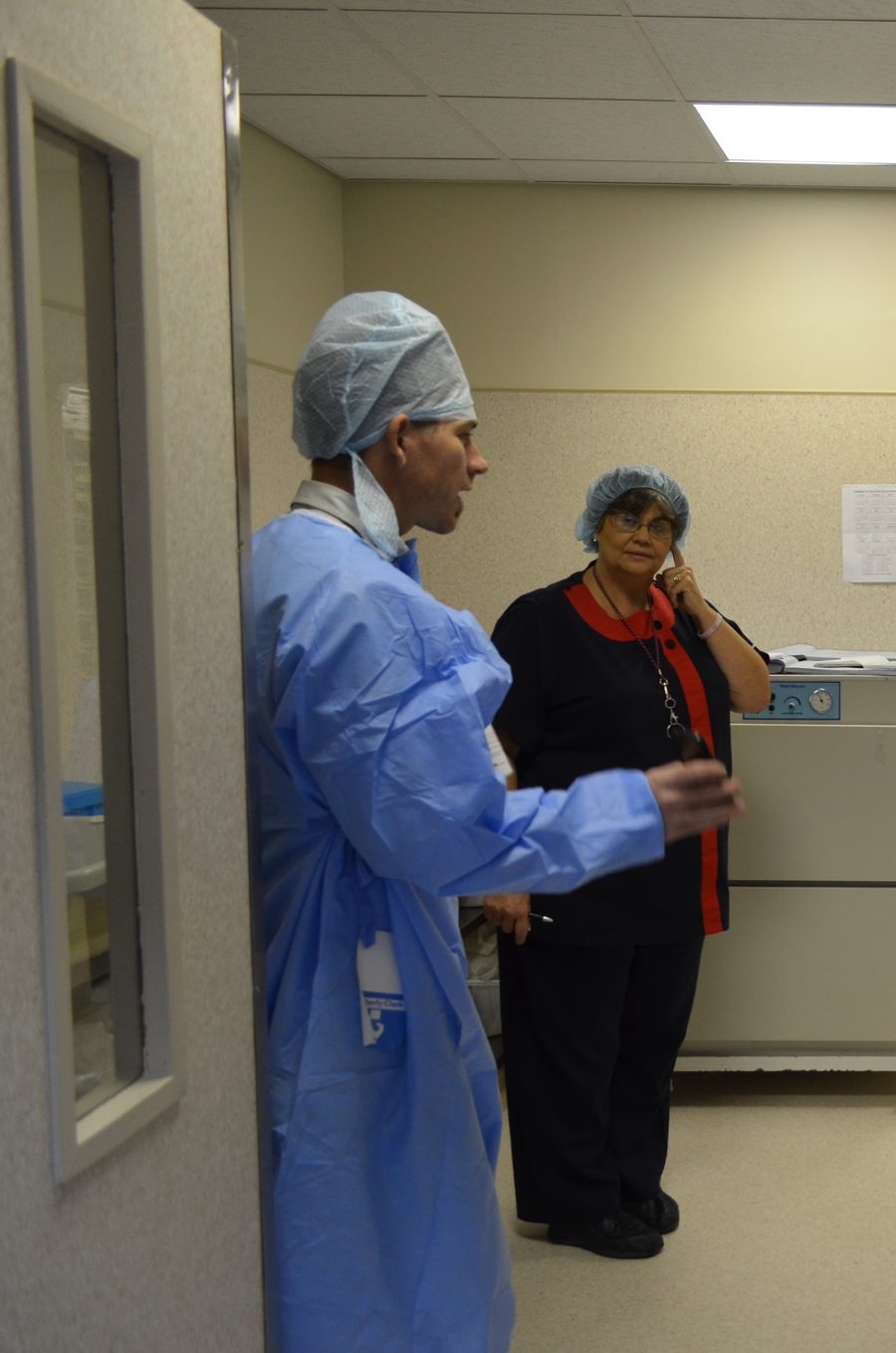 Staff in the ante room outside theatre, one gowned in blue, talking beside the shelving.