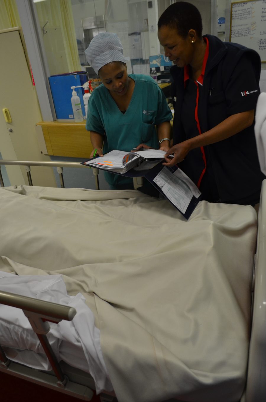 A nurse and a clinician reviewing a folder of documentation together at a patient's bedside.