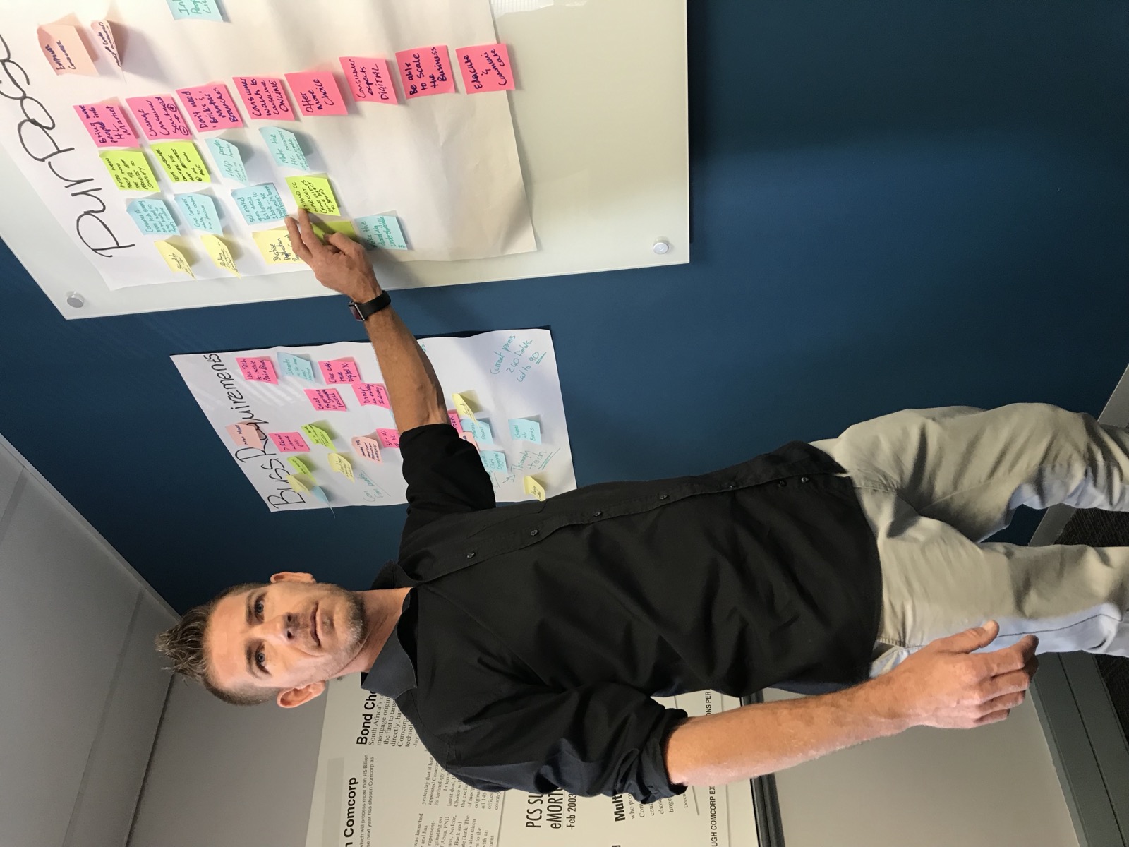 A man pointing at a Purpose whiteboard, with a printed article about Comcorp's mortgage origination business on the wall beside him.