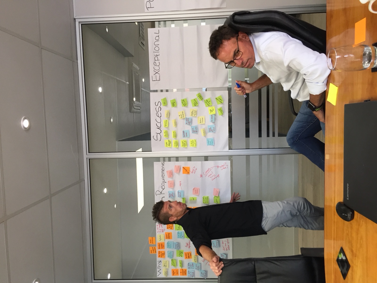 A man presenting Why, Business Requirements, Success and Exceptional whiteboards to a colleague seated at a boardroom table.